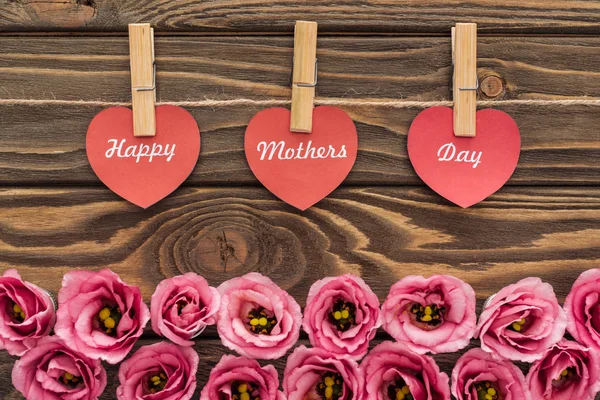 Vista dall'alto di fiori di eustoma rosa, mollette di vestiti e cuori di carta rossa con scritte di giorno di madri felici su tavolo di legno — Foto stock