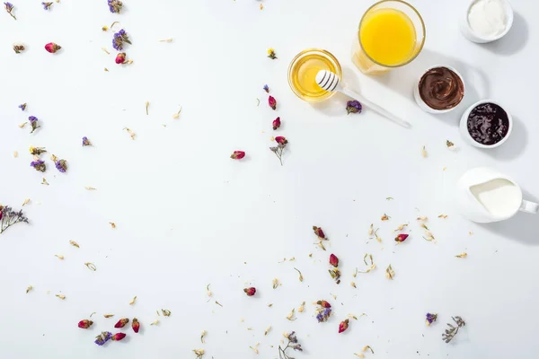 Vista dall'alto di ciotole con marmellata e crema al cioccolato vicino al miele, panna acida, bicchiere di succo d'arancia e brocca con latte su bianco — Foto stock