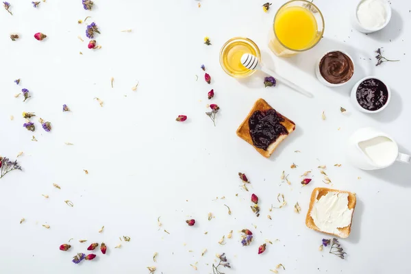 Top view of toasts with jam and cream cheese near honey, glass of orange juice and jug with milk on white — Stock Photo