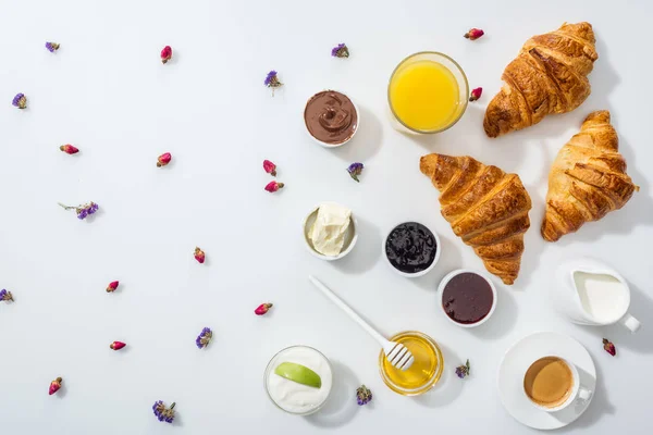 Vista dall'alto di gustosi croissant vicino a ciotole con marmellata, succo d'arancia e fiori secchi su bianco — Foto stock