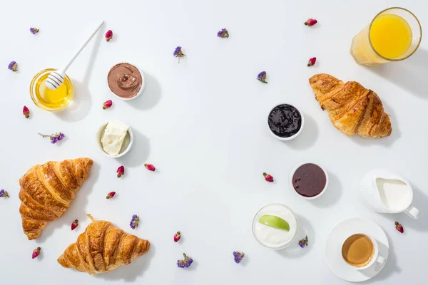 Vista dall'alto di cornetti vicino a bocce con marmellata, succo d'arancia e fiori secchi su bianco — Foto stock