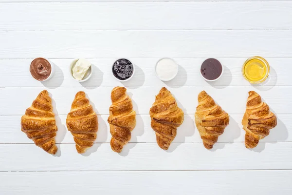 Top view of bowls with cream cheese, chocolate cream, sour cream, jam and honey near croissants on white — Stock Photo