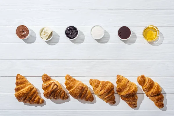 Vista superior de tigelas com creme de queijo, creme de chocolate, creme azedo, geléia e mel perto de croissants frescos em branco — Fotografia de Stock