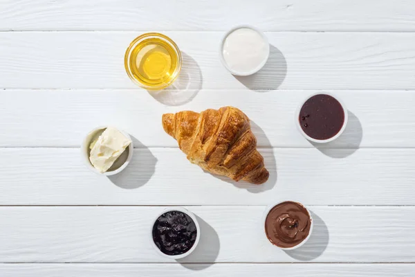 Flat lay of bowls with cream cheese, chocolate cream, sour cream, jam and honey near fresh croissant on white — Stock Photo