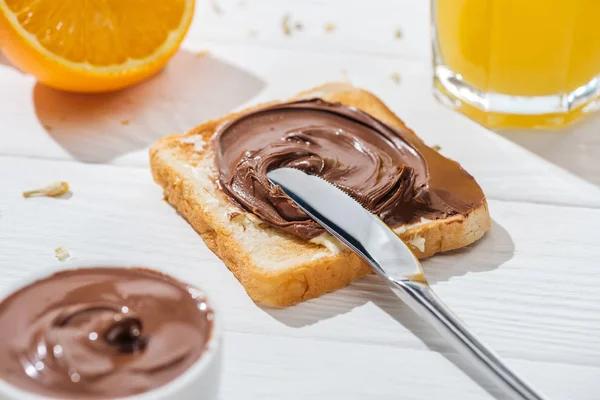 Fuoco selettivo di pane tostato con crema di cioccolato vicino a coltello e metà di arancione su bianco — Foto stock