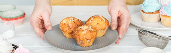 Panoramic shot of woman holding plate with muffins on table — Stock Photo