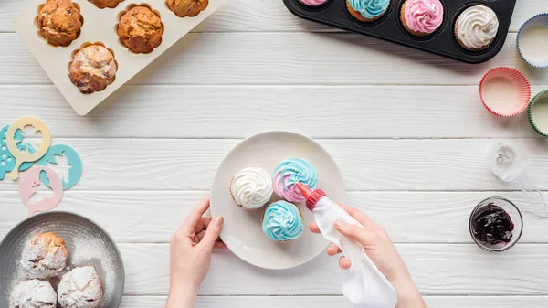 Partial view of woman decorating cupcakes with icing bag on table with cupcake trays — Stock Photo