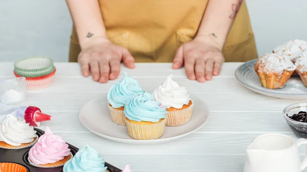 Vista ritagliata della donna vicino al tavolo con piatto con gustosi cupcake su grigio — Foto stock