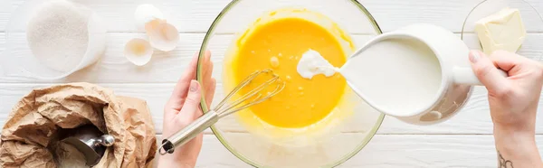 Tiro panorâmico de mulher derramando leite em massa líquida na mesa com ingredientes — Fotografia de Stock