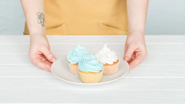 Cropped view of woman holding plate with delicious cupcakes on grey — Stock Photo