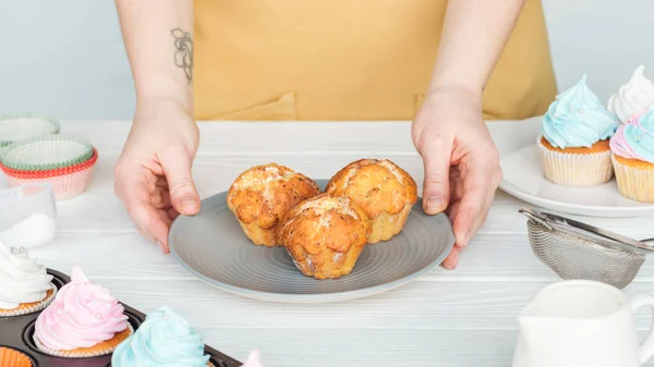 Cropped view of woman holding plate with cupcakes on wooden table isolated on grey — Stock Photo