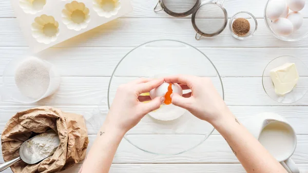 Частичный вид женщины, разбивающей яйцо в миске во время приготовления пищи на деревянном столе — стоковое фото