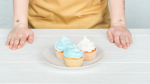 Vista cortada da mulher perto da placa com deliciosos cupcakes na mesa branca no cinza — Fotografia de Stock