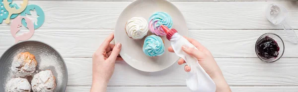 Panoramic shot of woman decorating cupcakes with icing bag on white table — Stock Photo