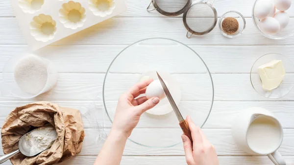 Vista recortada de la mujer con el cuchillo que agrieta el huevo en un tazón mientras se cocina en la mesa - foto de stock