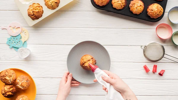 Cropped view of woman decorating cupcake with icing bag on table with cupcake trays — Stock Photo