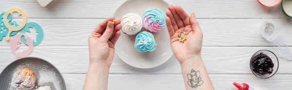 Panoramic shot of woman holding sprinkles while decorating cupcakes on white table — Stock Photo