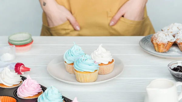 Vista cortada da mulher com as mãos nos bolsos perto da mesa com cupcakes isolados em cinza — Fotografia de Stock