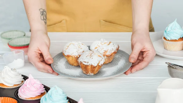 Vue recadrée de la femme tenant la plaque avec des cupcakes avec du sucre en poudre sur gris — Photo de stock