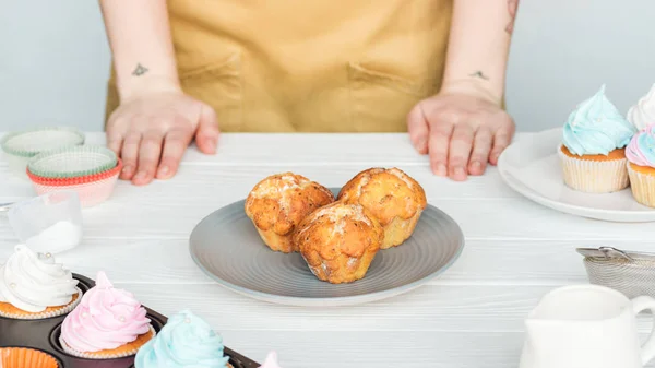Teilansicht einer Frau am Tisch mit Teller mit Cupcakes isoliert auf grau — Stockfoto