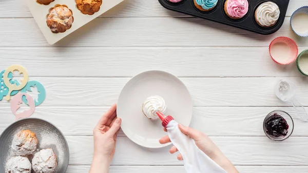 Cropped view of woman decorating cupcake with icing bag on table with cupcake trays — Stock Photo