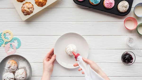 Vista ritagliata di donna decorazione cupcake con sacchetto di glassa sul tavolo con vassoi cupcake — Foto stock