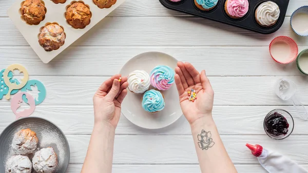 Cropped of woman holding sprinkles while decorating cupcakes on white table — Stock Photo