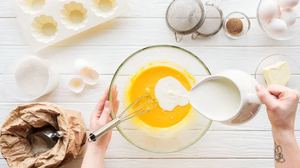 Vista recortada de la mujer vertiendo leche en masa líquida en la mesa con ingredientes - foto de stock