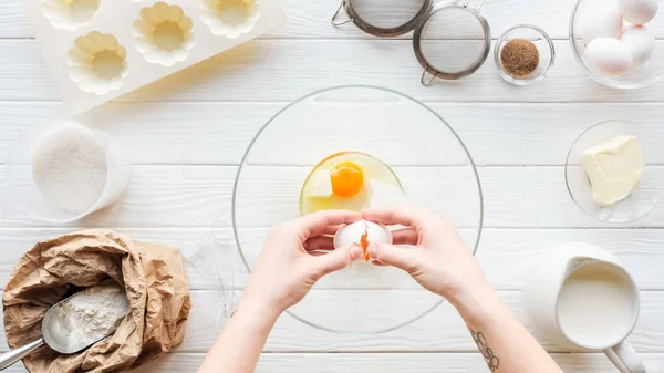 Vista recortada de la mujer agrietando huevo en un tazón mientras se cocina en la mesa - foto de stock