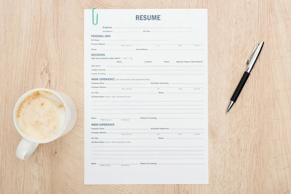 Top view of pen and cup of cappuccino with foam near resume template on wooden surface — Stock Photo