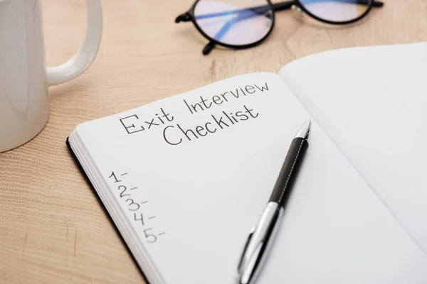 Notebook with exit interview checklist lettering and numbers on wooden table with pen, glasses and cup — Stock Photo