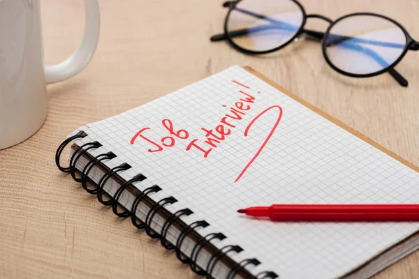 Caderno com entrevista de emprego lettering em mesa de madeira com marcador vermelho, óculos e copo — Fotografia de Stock