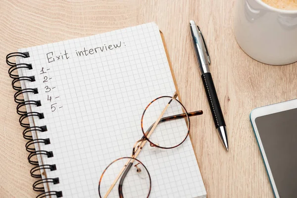 Notebook with exit interview lettering and numbers on wooden table near smartphone, pen, glasses and coffee cup — Stock Photo