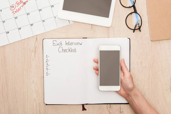 Vista recortada del reclutador que sostiene el teléfono inteligente con pantalla en blanco por encima del cuaderno con letras de lista de verificación de entrevista de salida - foto de stock