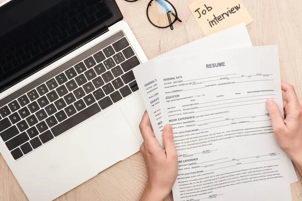 Cropped view of recruiter holding resume templates near sticky note with job interview lettering and laptop — Stock Photo