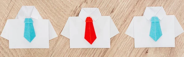 Panoramic shot of origami white shirts with blue ties with one red on wooden background — Stock Photo