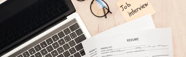 Panoramic shot of laptop, glasses, resume templates and sticky note with job interview lettering on wooden table — Stock Photo