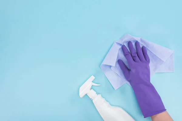 Обрізаний вид очищувача в фіолетовій гумовій рукавичці, що тримає ганчірку біля пляшки спрею з миючим засобом на синьому фоні — стокове фото
