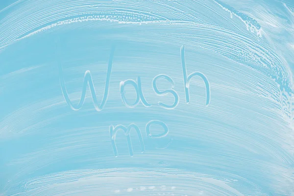 Wash me handwritten lettering written on glass with white foam on blue background — Stock Photo