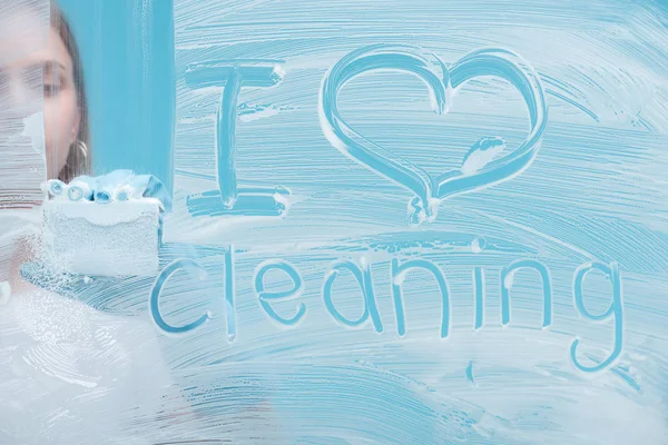 Частичный вид женщины, моющей стекло губкой рядом с надписью I love cleaning leing, написанной на белой пене на синем фоне — стоковое фото