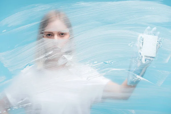 Селективный фокус женских глаз, смотрящих сквозь стекло с белой пеной на синем фоне — стоковое фото