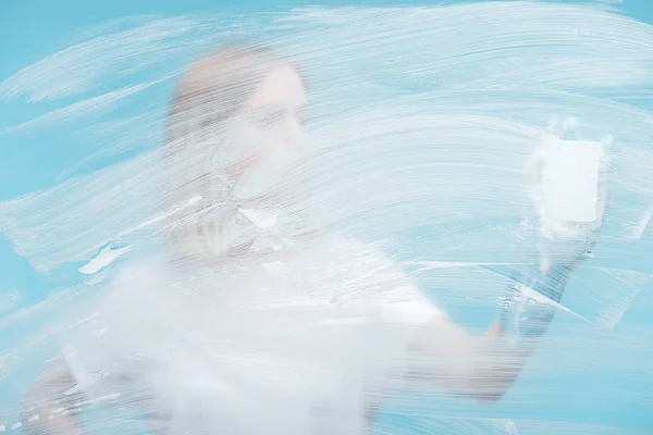 Mujer con esponja detrás de vidrio con espuma blanca sobre fondo azul - foto de stock