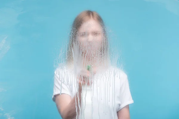 Selektiver Fokus einer jungen Frau, die Glas mit tropfendem Waschmittel auf blauem Hintergrund reinigt — Stockfoto