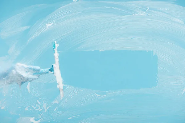 Cropped view of woman in rubber glove cleaning glass with squeegee on blue background — Stock Photo