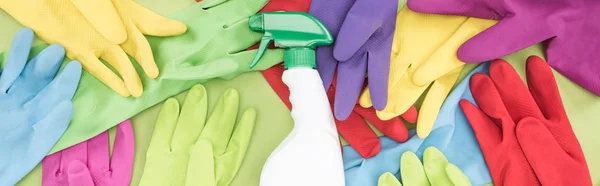 Panoramic shot of messy scattered multicolored rubber gloves and white spray bottle with cleanser on green background — Stock Photo