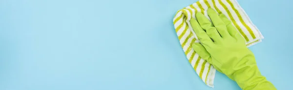 Panoramic shot of cleaner in green rubber glove holding striped rag on blue background — Stock Photo