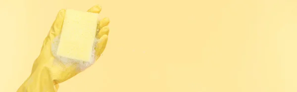 Plano panorámico de limpiador en guante de goma amarillo sosteniendo esponja con burbujas sobre fondo amarillo - foto de stock