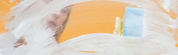 Panoramic shot of young woman in rubber glove with sponge cleaning glass covered with foam — Stock Photo