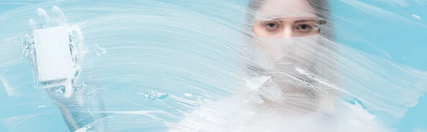 Панорамный снимок женских глаз, смотрящих через стекло с белой пеной на синем фоне — стоковое фото