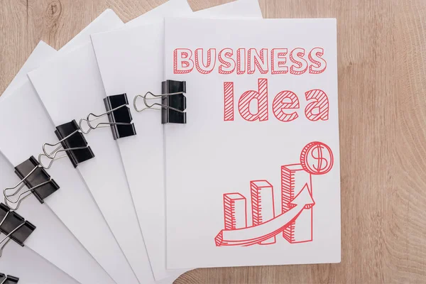 Vista dall'alto di fogli di carta bianca disposti con clip legante, iscrizione idea di business e illustrazione infografica su tavolo in legno, concetto di business — Foto stock